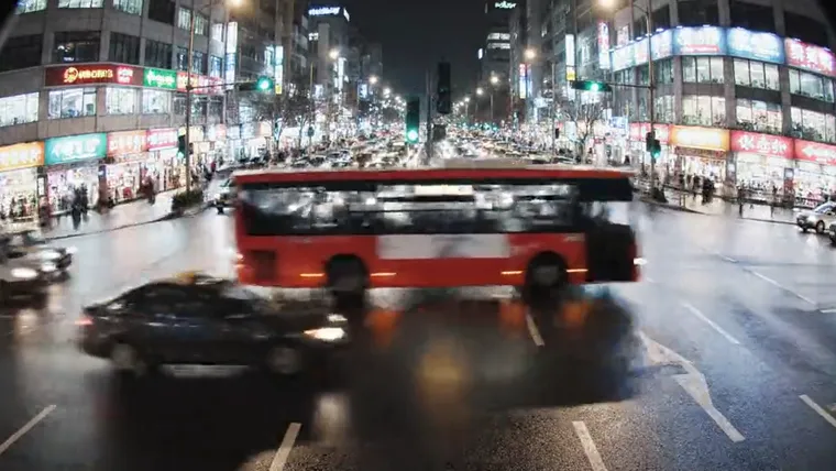 Night crossing on a live feed, a bus and cars mid-round under lit shopfronts